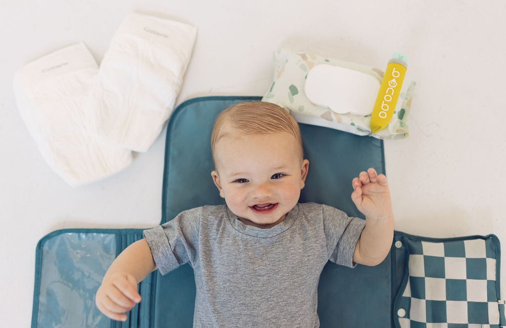Baby lying on a blue mat with a pack of wipes in the background that have the bonobo baby bidet secured to them.